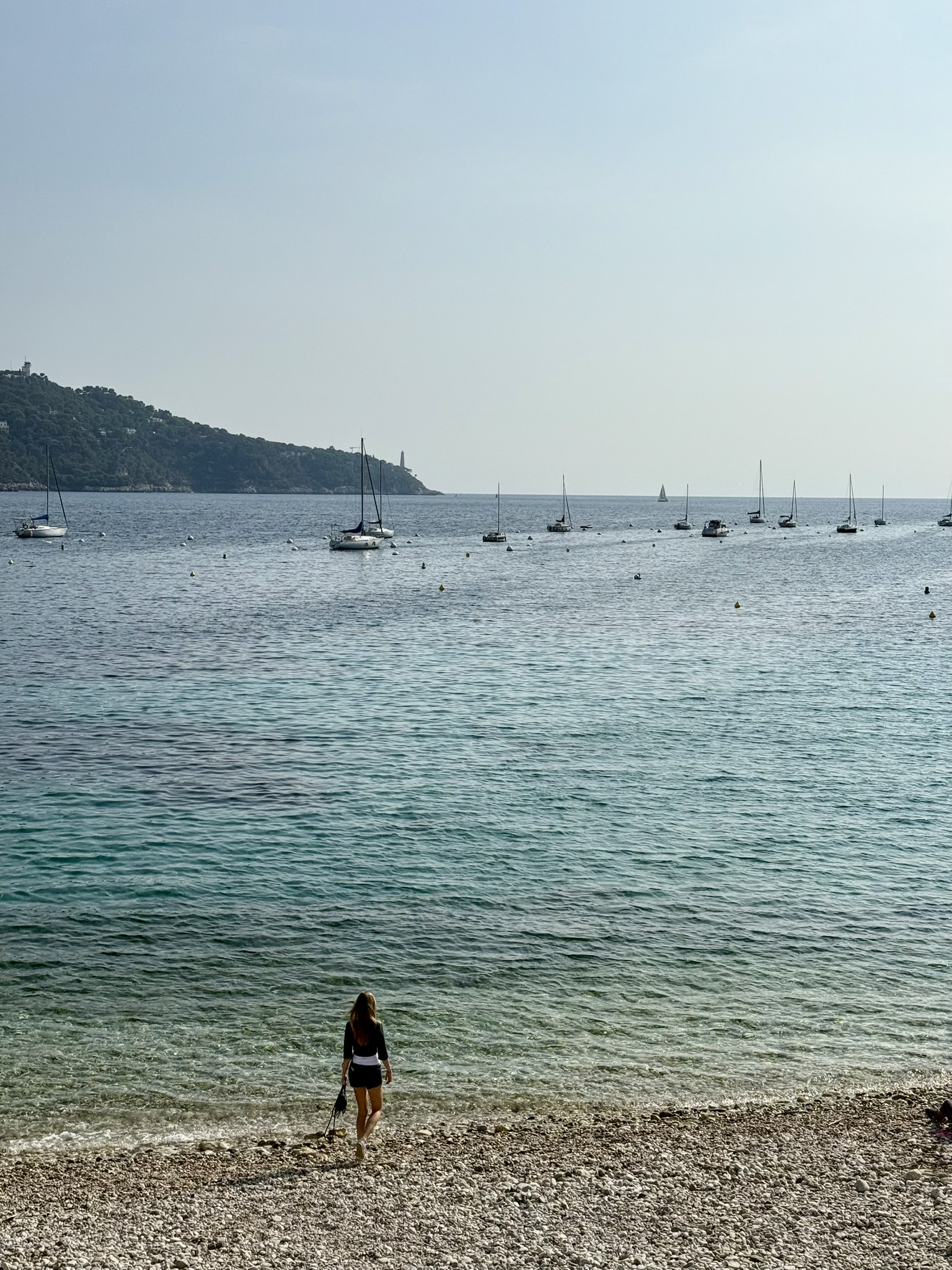 en tjej går på standen med utsikt över ett turkostblått Medelhav och segelbåtar som guppar på vattnet